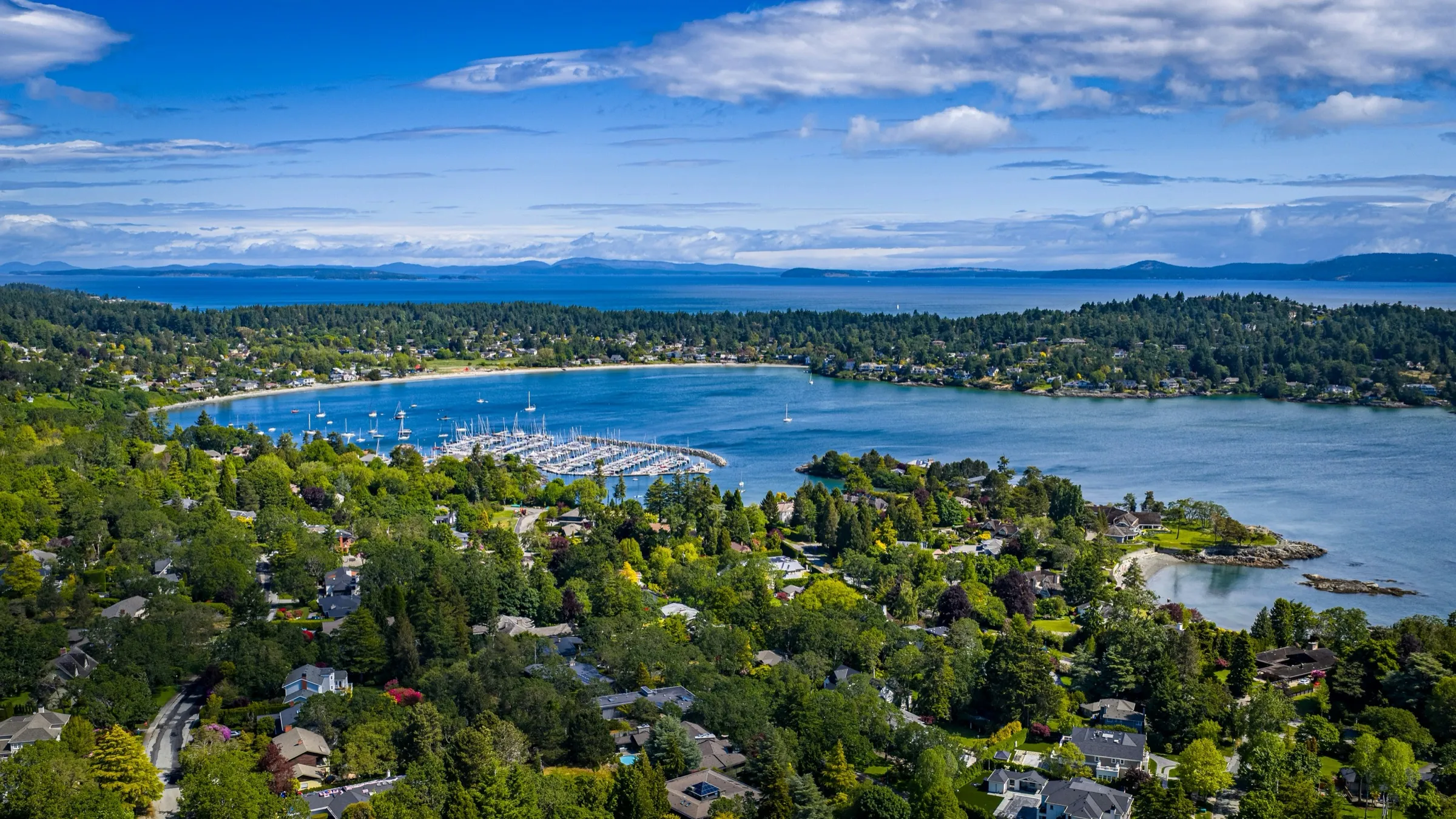 Aerial view of Cadboro Bay, with waterfront homes, wooded neighbourhoods and sailboats on the water